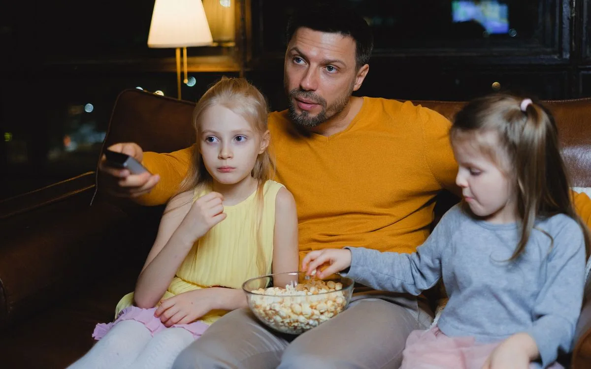 Un père et ses deux filles devant un film, un saladier de popcorn sur les genoux